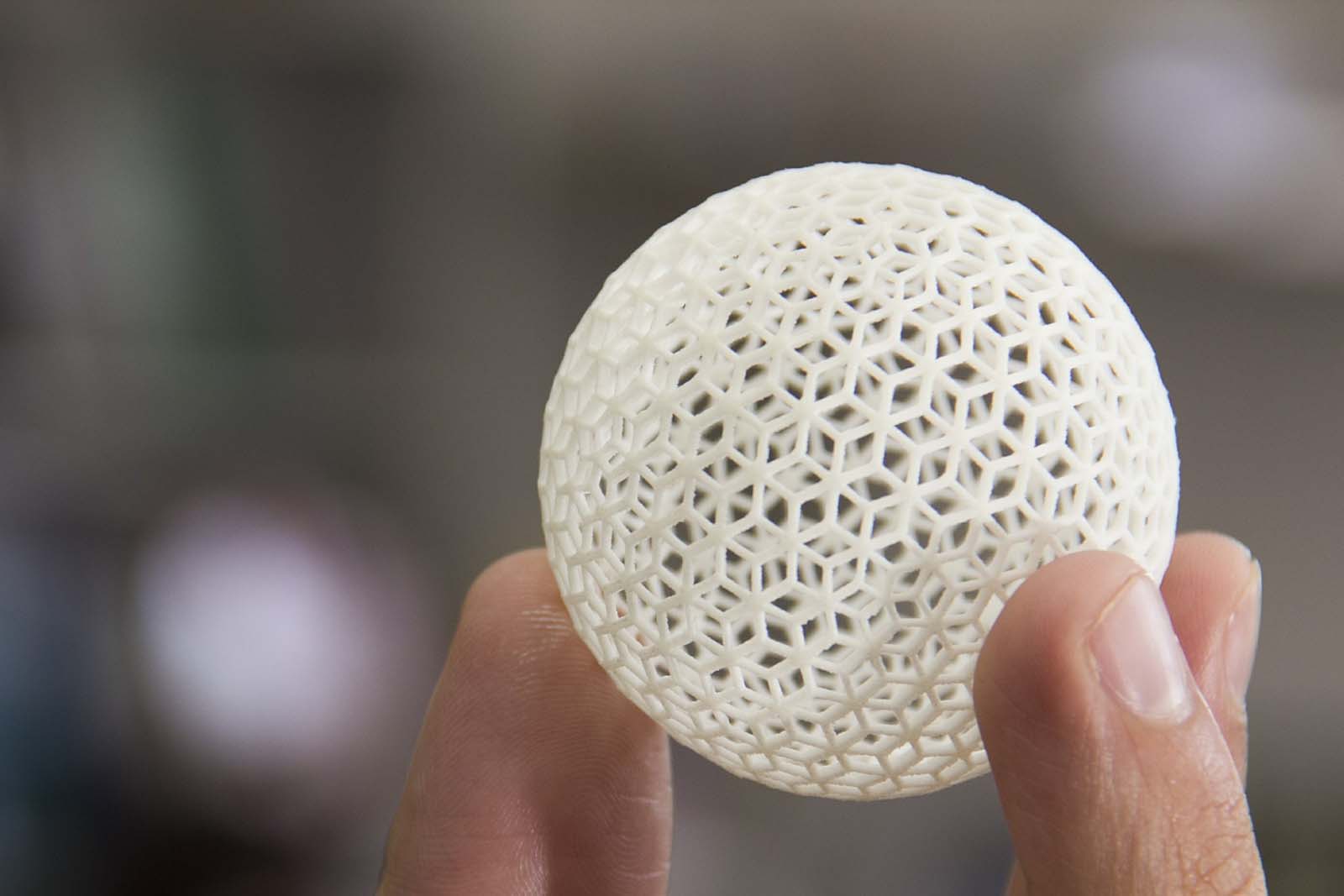 A hand holds a white, 3D-printed spherical object with an intricate hexagonal lattice structure.